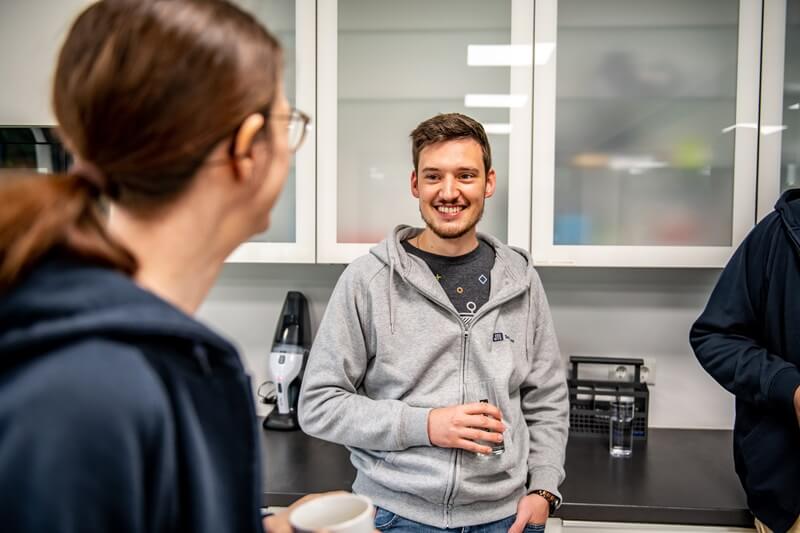 Drei Personen unterhalten sich in einer Büroküche. Ein Mann in grauer Kapuzenjacke mit dem Firmenlogo von JTL-Software hält ein Glas Wasser und lächelt. Eine Frau mit Brille und eine weitere Person sind teilweise sichtbar. Im Hintergrund stehen Küchenschränke, eine Kaffeemaschine und Gläser.