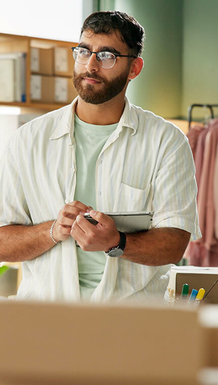 Ein junger Mann mit Bart, Brille und dunklem Haar steht in einem Lager oder Versandbereich. Er trägt ein helles Hemd über einem grünen T-Shirt und hält ein Tablet sowie einen Stift in den Händen. Im Hintergrund sind Kartons, Regale mit Lagerware und eine Kleiderstange mit hängender Kleidung zu sehen.