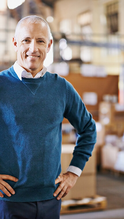 Ein älterer, lächelnder Mann mit kurz geschorenem, grauem Haar steht mit den Händen in die Hüften gestützt in einem Lager oder einer Produktionshalle. Er trägt ein weißes Hemd unter einem blauen Pullover. Im Hintergrund sind gestapelte Kartons und Regale mit unscharfen Details zu sehen.