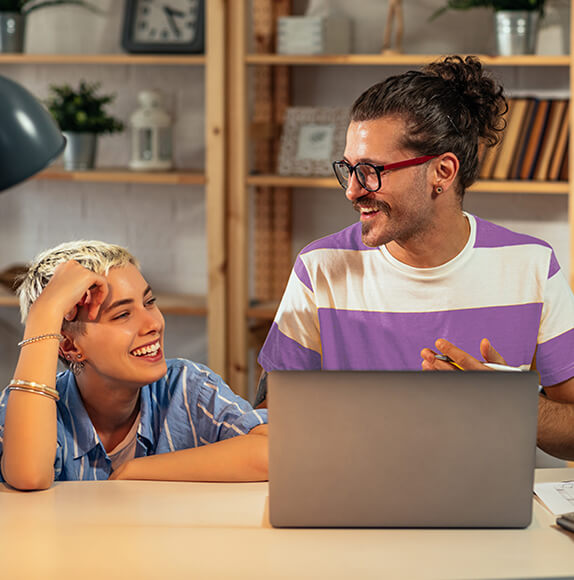 Zwei junge Menschen sitzen lachend an einem Tisch mit einem Laptop. Eine Person hat kurze, blond gefärbte Haare, die andere trägt einen Dutt und eine Brille. Im Hintergrund stehen Holzregale mit Büchern, Deko und einer Uhr.